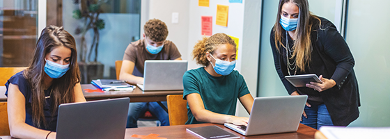 Teacher and students wearing masks using laptops and tablets in a socially distanced classroom setting