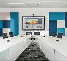 Modern conference room with video display, conferencing system, and white tables arranged in a U‑shape
