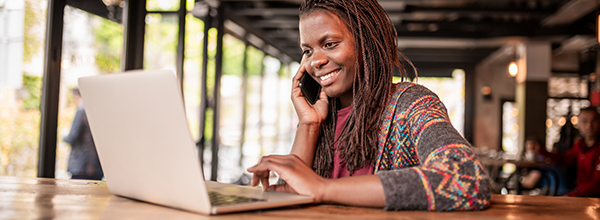 Woman working remotely in a café, smiling while talking on smartphone and using laptop