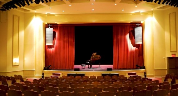 Empty theater with red curtains, grand piano on stage, and line array speakers for live performances