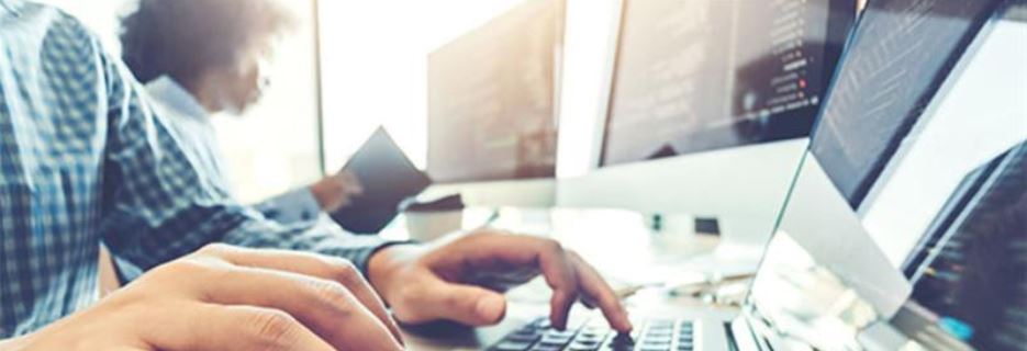 Close-up of hands typing on laptop in modern office with multiple monitors and coworker in background