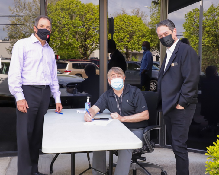 Three men wearing masks gather around an outdoor check‑in table, one seated and signing paperwork.