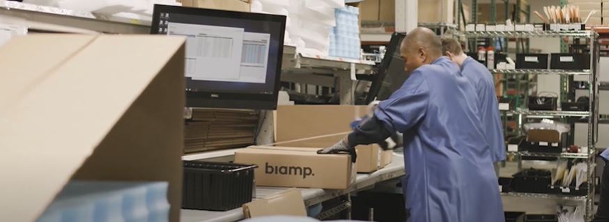 Two warehouse workers in blue coats pack a Biamp box for shipment on a production line.