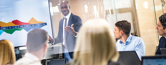 Business meeting where a presenter gestures at a screen with colorful charts while colleagues listen around a table.