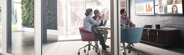 Three people in a modern conference room conduct a video meeting, looking at two screens showing a presentation and remote attendees.