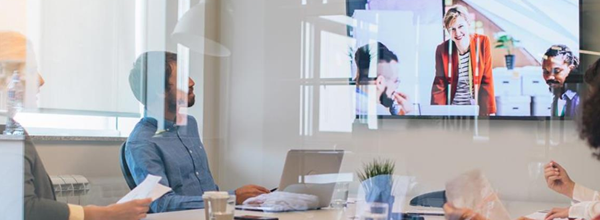 A group of people sit at a conference table, looking up at a large screen displaying a video conference call.
