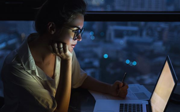 A woman in glasses works at her laptop late at night, with the screen illuminating her face as she writes.