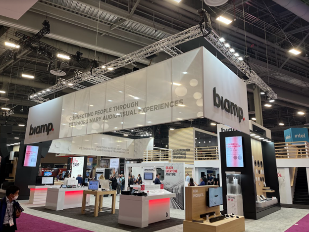 Biamp trade show booth with illuminated hanging sign, product demo stations, and messaging about connecting people through extraordinary audiovisual experiences.