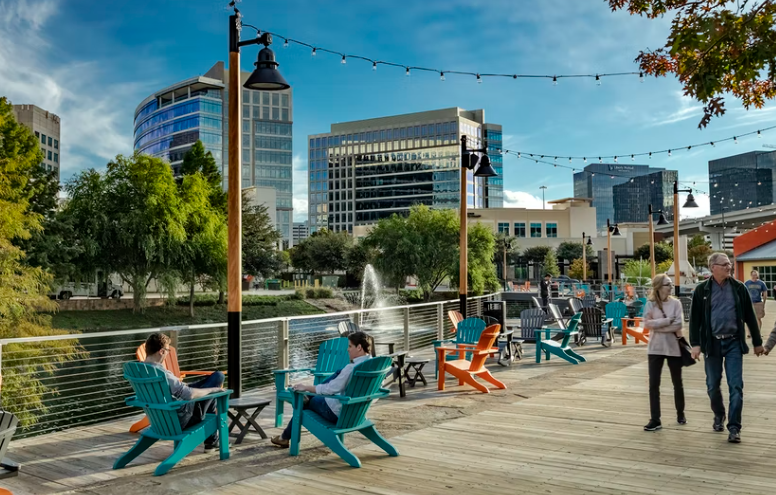 Scenic urban boardwalk with string lights, colorful chairs, and people relaxing along a waterfront in a modern downtown business district.