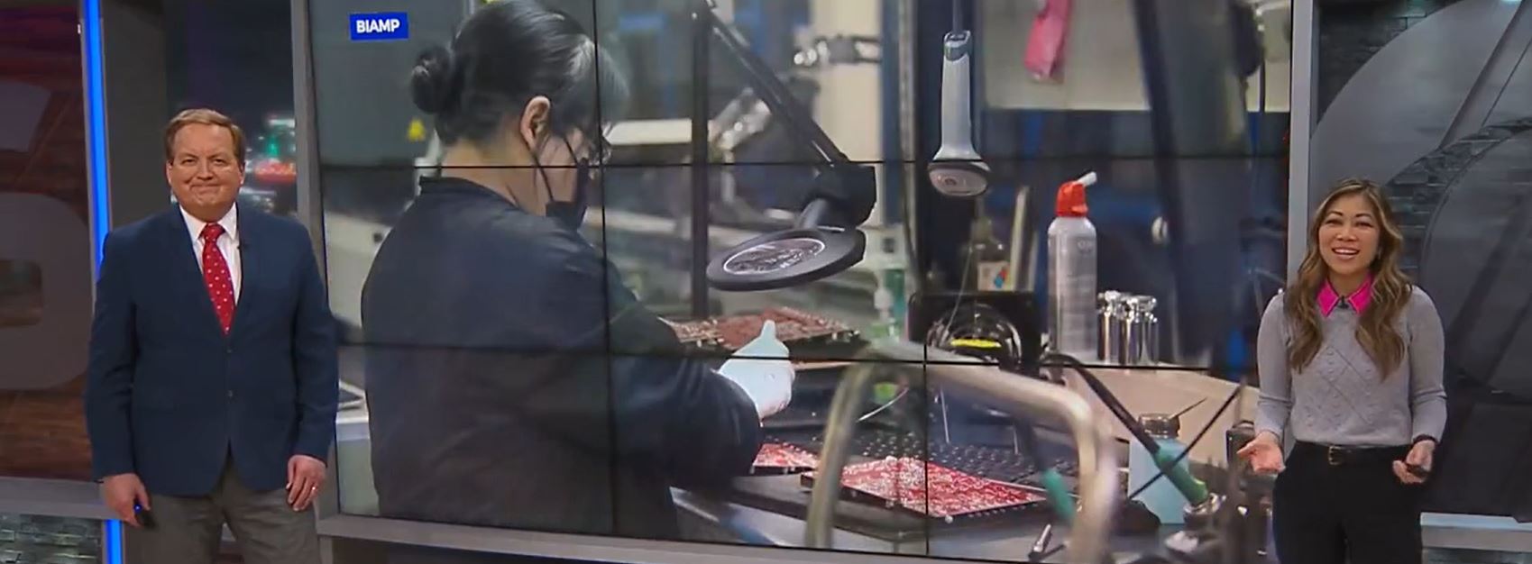 Television news segment with two anchors standing beside large video wall showing Biamp manufacturing employee working on electronics assembly.