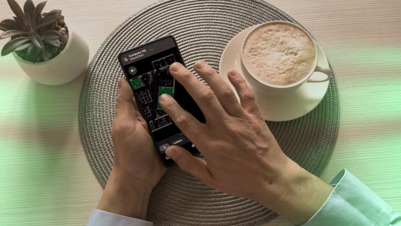 Person using a smartphone workplace app to view an office floor plan while sitting at a table with coffee, showing mobile room booking.