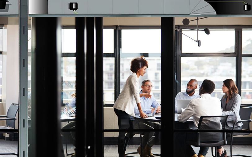 Modern glass-walled conference room with diverse team collaborating around a table, illustrating technology-enabled workplace meetings and communication.