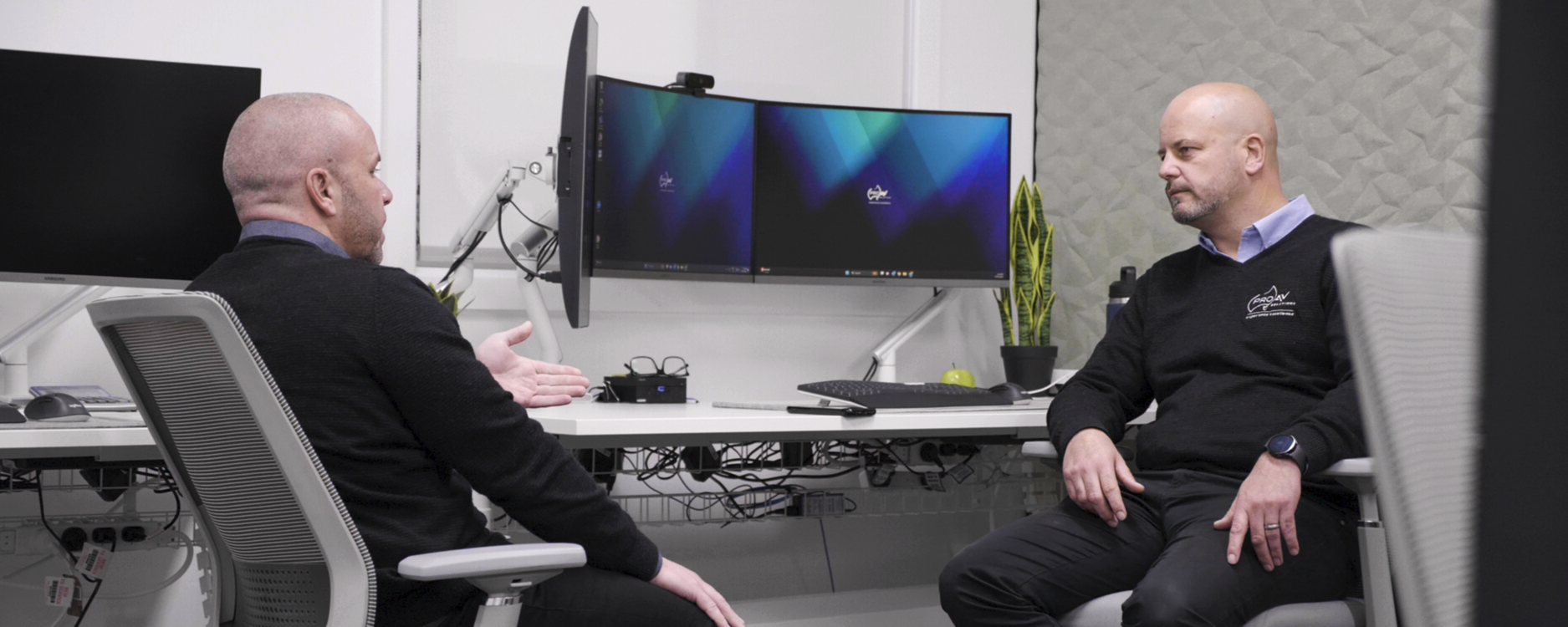 Two professionals seated in a modern office having a discussion in front of dual monitors, illustrating collaborative workplace consultation.