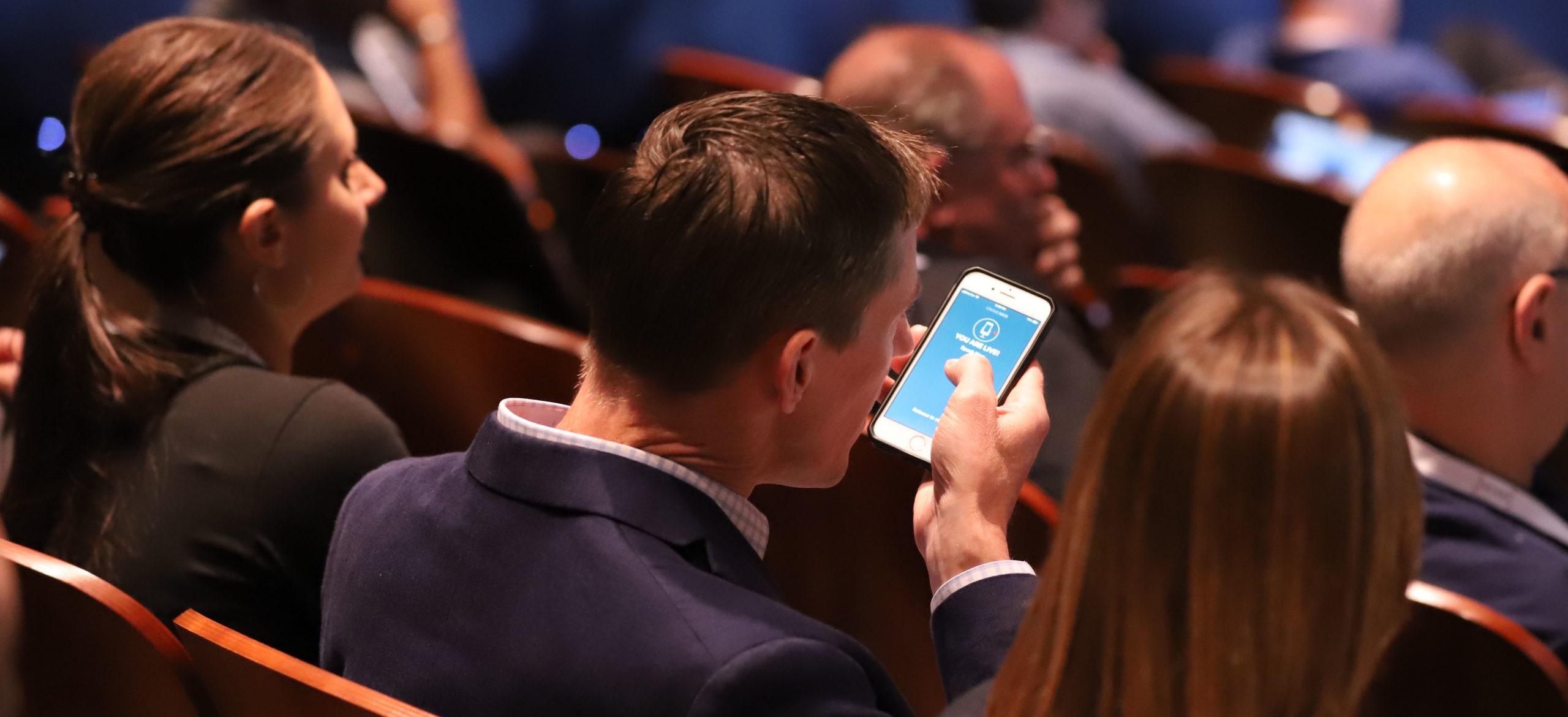 Audience member using smartphone app during live presentation in auditorium, illustrating interactive participation and mobile event engagement tools.