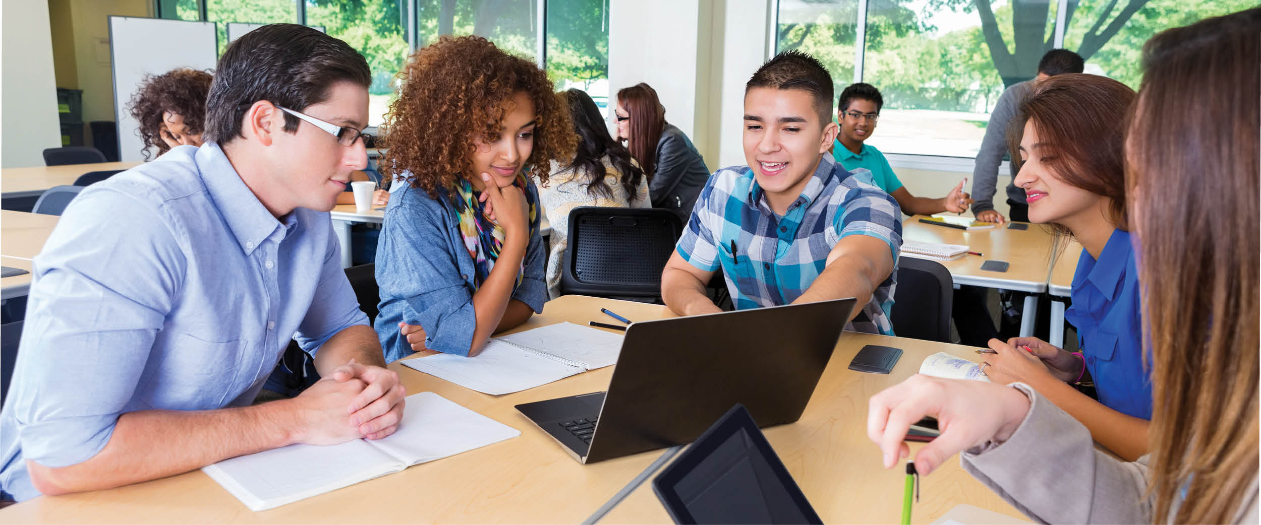 College students collaborating in an active learning classroom, working together around a laptop and digital devices on shared tables.
