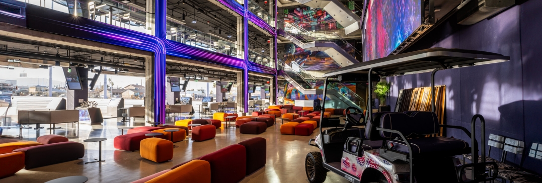 Vibrant modern atrium space with colorful seating, neon lighting, large digital screens and a golf cart in the foreground.