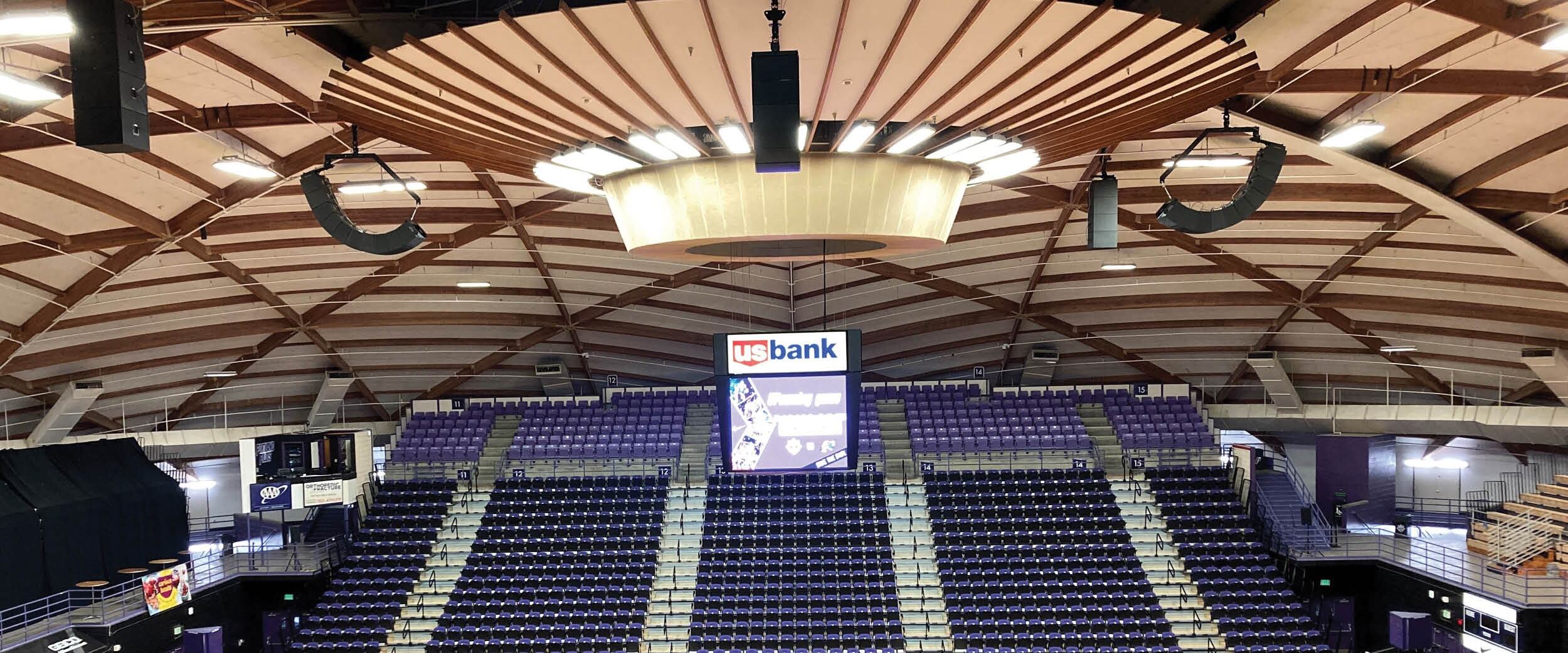 Indoor sports arena with purple seating, central scoreboard, and suspended loudspeaker arrays under distinctive wood‑beam dome roof.