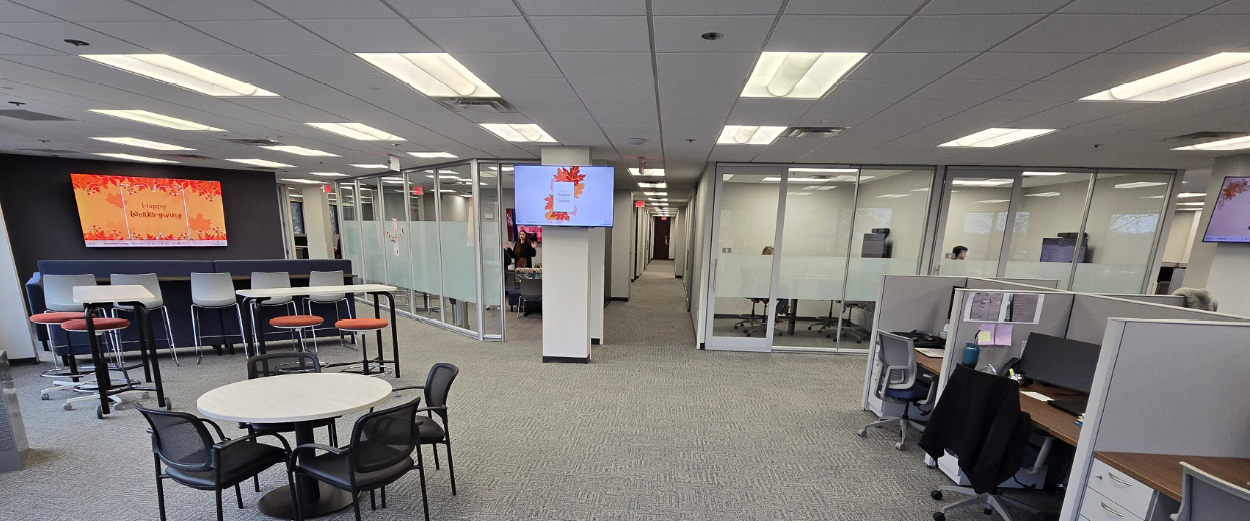 Open-plan modern office with glass-walled meeting rooms, digital signage displays, collaboration tables, and cubicle workstations along the corridor.