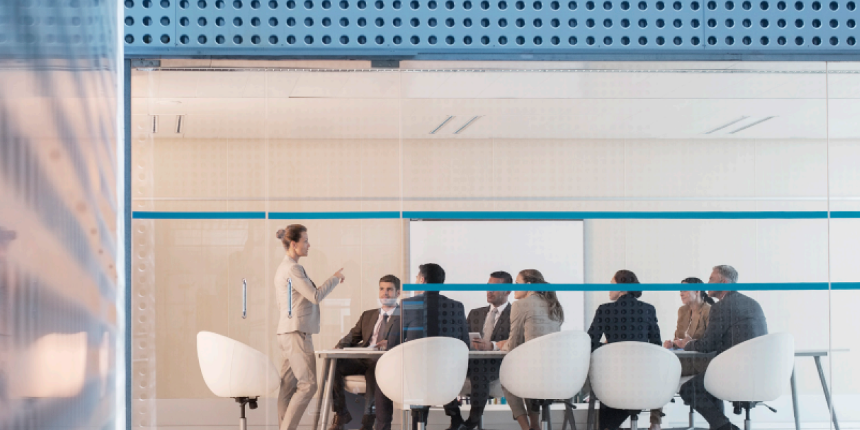 Modern glass-walled conference room with business professionals seated around a table listening to a presenter.
