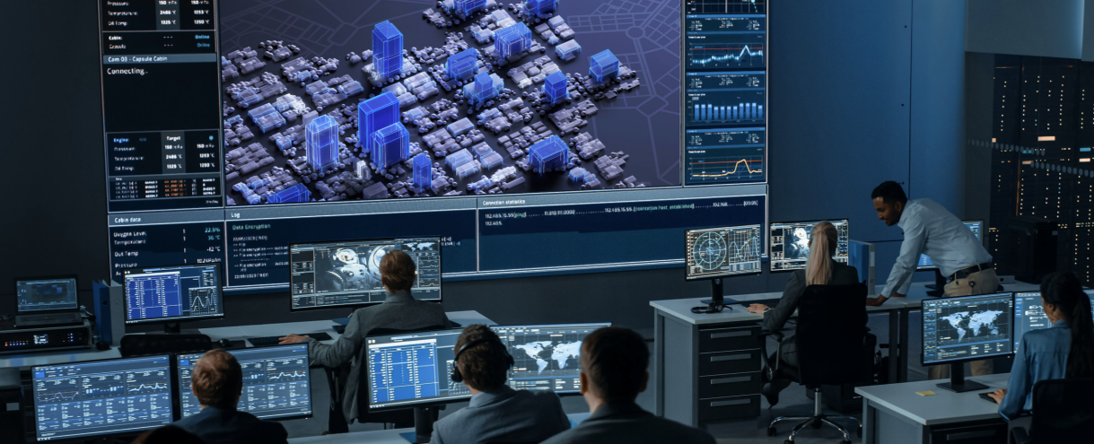 High-tech command center with analysts at computer workstations monitoring data and a large wall display showing a 3D city map and dashboards.