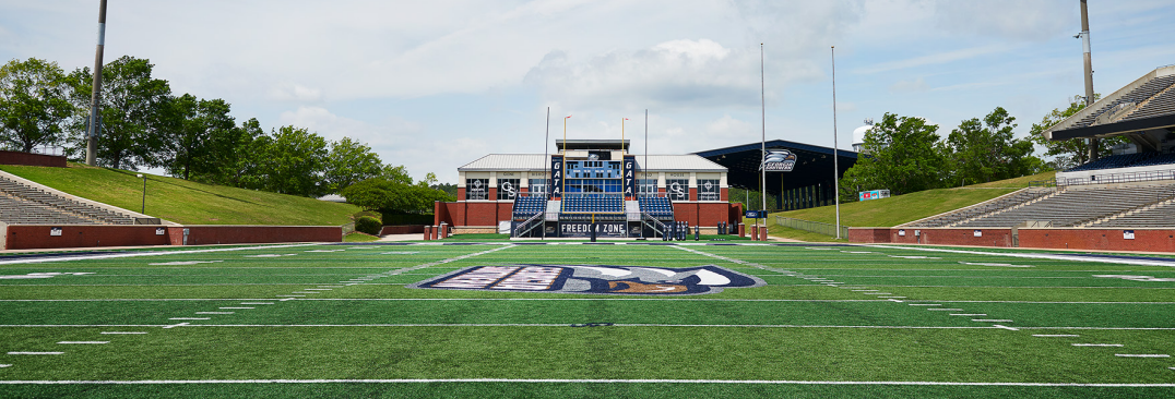 Wide-angle view of an empty American football stadium field with logo at midfield and bleachers on both sides.